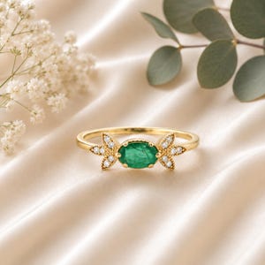 May include: A gold ring featuring a central oval-cut green gemstone surrounded by a delicate milgrain edge. The band is accented on both sides by a cluster of three small, round, clear white diamonds arranged in a leaf-like pattern.