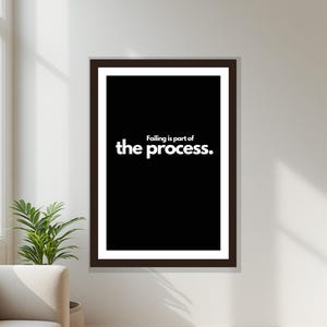 May include: A framed print with the text "Failing is part of the process" in white on a black background. The print is in a brown frame and hangs on a white wall. A potted plant and a beige sofa are visible in the corner.