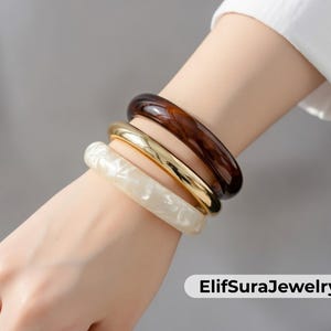 Puede incluir: Cuatro brazaletes apilados en tonos marrón, dorado y blanco. El brazalete superior es marrón oscuro, seguido de uno dorado y otro blanco veteado. El brazalete inferior es de color crema claro. Las joyas están en la muñeca de una persona.