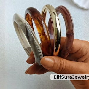 Pode incluir: Uma mão segura uma coleção de pulseiras em várias cores. As pulseiras incluem uma branca e cinza marmorizada, um padrão de tartaruga, uma dourada e uma bordô escura. As joias são lisas e brilhantes.