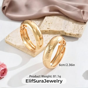 May include: Two gold-colored bangles, one with a smooth finish and the other with a hammered texture, are displayed on a light-colored surface. The bangles are 6 inches in diameter. Pink roses and a silk cloth are in the background.