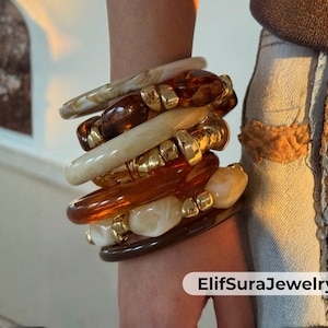 以下が含まれることがあります： 茶色、クリーム色、金色のブレスレットのスタック。 ブレスレットは、滑らかなバングルやビーズのデザインなど、さまざまな形と質感を持っています。 金色の金属アクセントが、ブレスレットの間から見えます。 ブレスレットは手首に重ねられています。