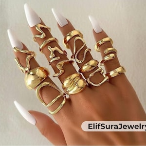 May include: A hand displays a collection of gold-colored rings in various designs. The rings include smooth bands, abstract shapes, and open designs. The rings are worn on multiple fingers, showcasing the variety of styles. The image includes the text "ElifSuraJewelry".