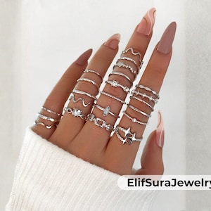 May include: A selection of silver rings in diverse styles, such as wavy, chain-link, and star designs. Some rings are adorned with small gemstones. The rings are shown on a hand with long, manicured nails. The brand name "ElifSuraJewelry" is visible.