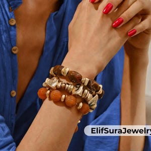 Könnte beinhalten: Drei gestapelte Armbänder mit braunen, orangefarbenen und silbernen Perlen. Die Armbänder werden an einem Handgelenk mit rotem Nagellack getragen. Der Hintergrund ist ein blaues Hemd. Der Text "ElifSuraJewelry" ist sichtbar.