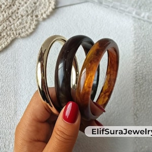Könnte beinhalten: Drei Armreifen in Gold-, Dunkelbraun- und Bernsteinfarben. Der goldene Armreif hat ein glänzendes Finish, während die anderen beiden glänzend aussehen. Die Armreifen werden von einer Hand mit rotem Nagellack gehalten. Der Markenname "ElifSuraJewelry" ist sichtbar.