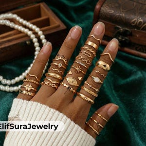 May include: A hand displaying numerous gold rings in diverse styles, such as plain bands, twisted designs, and geometric patterns. Some rings incorporate small details like a star or a gemstone. The rings are set against a white sweater and a green fabric background.