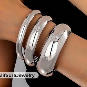 May include: A close-up of a silver-colored bracelet with three thick, rounded bands. The shiny metal reflects light, creating highlights and shadows. The jewelry is worn on a wrist, with the brand name "ElifSuraJewelry" visible.