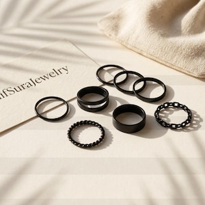 May include: A set of seven black rings in various designs, including smooth bands, a double-band ring, a twisted ring, and a chain-link ring. A white card with the text ElifSuraJewelry is positioned next to the rings and a small fabric pouch.