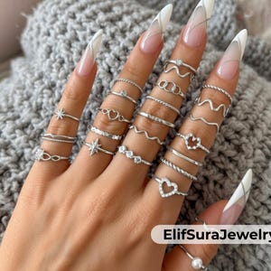 Può includere: Una collezione di anelli d'argento con vari design, tra cui stelle, cuori e motivi ondulati. Gli anelli sono esposti su una mano con unghie lunghe e curate. Gli anelli sono delicati ed eleganti, perfetti per l'uso quotidiano.
