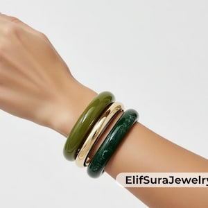 May include: Three bangles in olive green, gold, and dark green are stacked on a wrist. The bangles are smooth and glossy, with a rounded shape. The jewelry is displayed against a white background. The text "ElifSuraJewelry" is in the bottom right corner.