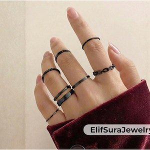May include: A hand wearing several black rings of various designs. The rings include a simple band, a chain link style, and textured bands. The rings are displayed against a burgundy fabric background. The text "ElifSuraJewelry" is visible.