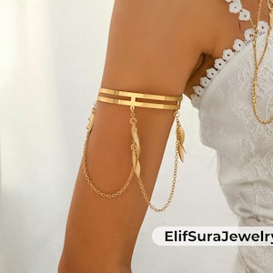 May include: A gold-coloured arm cuff with a double-band design, featuring dangling leaf charms and delicate chain accents. The jewellery piece is displayed on an arm, showcasing its elegant and bohemian style. The jewellery is from ElifSuraJewelry.