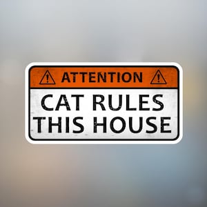 May include: A rectangular sign with a black border and a white background. The top section is orange with the word "ATTENTION" and an exclamation point symbol. The text "CAT RULES THIS HOUSE" is in bold black font.