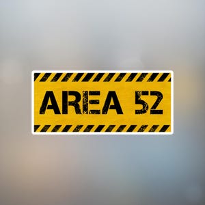 May include: A rectangular sticker with the text "AREA 52" in bold black letters on a yellow background. The sticker has black and yellow diagonal stripes along the top and bottom edges, with a white border.