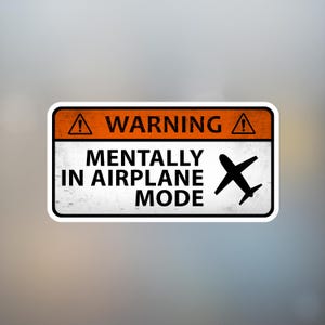 Può includere: Un adesivo rettangolare con sfondo bianco e bordo nero. La sezione superiore è arancione con la parola "WARNING" in bianco. Sotto, il testo recita "MENTALLY IN AIRPLANE MODE" con una grafica di aereo nero.