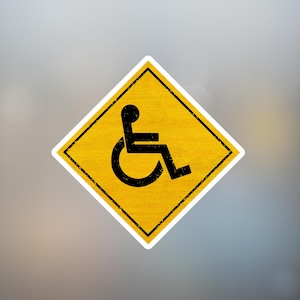 May include: A yellow diamond-shaped sign with a black wheelchair symbol. The sign has a black border and a textured appearance. The background is a blurred gradient of gray and white.