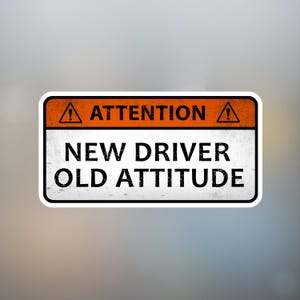 May include: A rectangular sticker with a white background and black text. The top section is orange with the word "ATTENTION" and a warning symbol. The text below reads "NEW DRIVER OLD ATTITUDE".