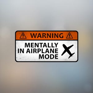 May include: A rectangular sticker with a black border and a blurred background. The sticker has an orange top section with the word "WARNING" and a warning symbol. Below, it reads "MENTALLY IN AIRPLANE MODE" with a black airplane graphic.