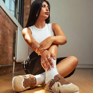Può includere: Un paio di zoccoli in camoscio color marrone e beige con punta chiusa, un ampio cinturino regolabile sul collo del piede con fibbia in metallo, dettagli bianchi e una spessa suola bianca, indossati con calzini bianchi.