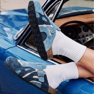 Könnte beinhalten: Ein Paar Clogs aus blauem Wildleder mit einem abstrakten Muster in verschiedenen Blau- und Cremetönen, einer Kork-Zwischensohle, einer schwarzen, strukturierten Gummilaufsohle und einem verstellbaren Metall-Schnallenriemen, getragen mit weißen Socken.