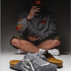 Könnte beinhalten: Ein Paar graue Wildleder-Clogs zum Hineinschlüpfen mit weißem, abstraktem Überlagerungsmuster, einem Riemen mit Metallschnalle, einem Fußbett in Korkoptik und einer schwarzen Gummilaufsohle. Das Schuhwerk hat eine runde Kappe und offene Ferse.