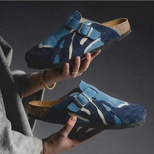 Puede incluir: Zuecos de ante azul con cierre de hebilla. Los zuecos presentan un diseño azul de varios tonos con detalles en blanco y una suela de corcho. Los zapatos se muestran sobre un fondo oscuro.
