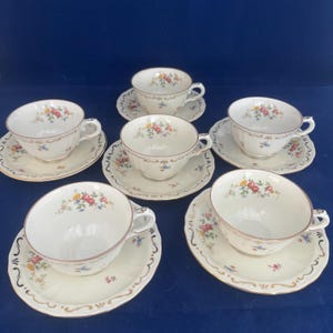 May include: Six white teacups and saucers with floral patterns. The cups and saucers have a scalloped edge and a gold trim. The floral pattern includes red, yellow, and blue flowers. The teacups are arranged on a dark blue surface.