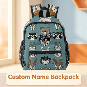 Puede incluir: Una mochila de color verde azulado con un patrón repetitivo de animales del bosque en dibujos animados, incluyendo osos, mapaches, búhos, erizos y ciervos. El bolsillo frontal muestra el nombre KAI en letras negras. Cremalleras y bolsillos de malla.