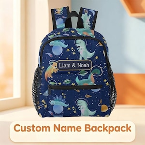 May include: A navy blue backpack featuring a repeating pattern of dinosaurs in space helmets, planets, and stars. The front pocket displays a rectangular label with the text Liam & Noah. The bag includes black straps and side mesh pockets.