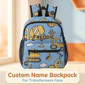May include: A light blue backpack featuring a repeating pattern of yellow construction vehicles, orange traffic cones, and white clouds. The front pocket displays the name FREDDIE in black capital letters centered on a cement mixer truck.