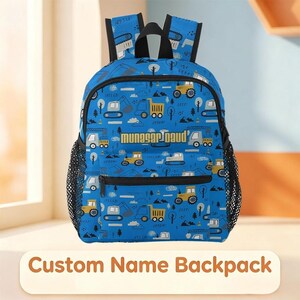 May include: A blue backpack featuring a repeating pattern of yellow and gray construction vehicles, trees, and clouds. The bag includes black shoulder straps, a front zippered pocket, and black mesh side pockets. The text "munasaraud" is printed in gold.