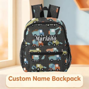 May include: A dark gray backpack featuring a repeating pattern of construction vehicles including excavators and dump trucks in shades of blue, orange, and green. The name Mariano is printed in white text on the front pocket, with black mesh side pockets.