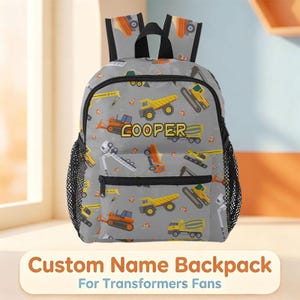 May include: A gray backpack featuring a repeating pattern of yellow and orange construction vehicles, including dump trucks and excavators, with orange traffic cones. The name COOPER is printed in yellow block letters on the front pocket.