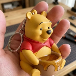 以下が含まれることがあります： 赤いシャツと蜂蜜の壺を持った、黄色のくまのプーさんのキーホルダーフィギュア。フィギュアは、滴る蜂蜜が入った茶色の壺を持っています。キーホルダーは丈夫な素材でできており、コレクターズアイテムです。