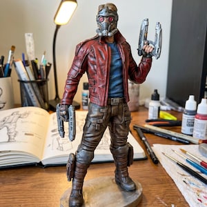 Op de afbeelding: Gedetailleerd beeldje van een personage in een rode leren jas, bruine broek en een masker, met twee zilveren straalkanonnen. Het beeldje staat op een kleine, ovale basis. De achtergrond omvat kunstbenodigdheden en een schetsboek.
