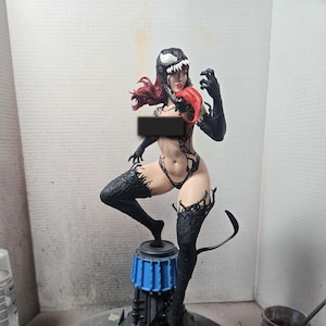 Op de afbeelding: Gedetailleerd beeldje van een vrouwelijk personage in een zwart-wit Venom-ge&iuml;nspireerd kostuum. De figuur heeft rood haar, een rode tong en zwarte details op de benen en armen. De figuur staat op een blauw-zwarte basis.