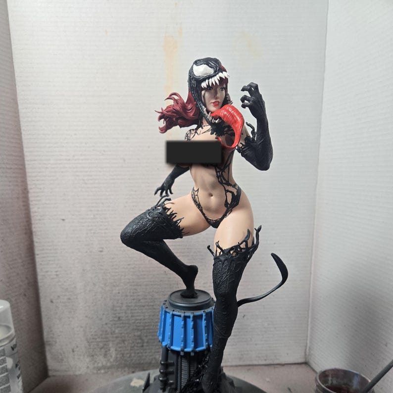 Op de afbeelding: Gedetailleerd beeldje van een vrouwelijk personage in een zwart-wit Venom-ge&iuml;nspireerd kostuum. De figuur heeft rood haar, een rode tong en zwarte details op de benen en armen. De figuur staat op een blauw-zwarte basis.