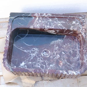 Peut inclure: Un lavabo rectangulaire sculpté dans du marbre brun rougeâtre foncé avec des veines blanches marquées. La vasque présente une surface intérieure lisse et polie avec un trou d'évacuation central, tandis que les côtés extérieurs sont cannelés.