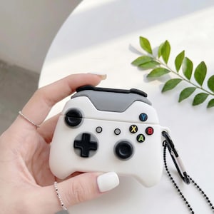 May include: White and gray controller-shaped case for wireless earbuds. The case features a black directional pad, buttons, and a gray top section. Colored buttons include blue, green, red, and yellow. A black chain and clasp are attached.