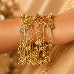 Könnte beinhalten: Eine Sammlung goldfarbener Armbänder schmückt ein Handgelenk. Jedes Armband hat eine Reihe von hängenden, mit Perlen besetzten Anhängern. Die Armbänder werden vor einem Hintergrund aus goldfarbenem Stoff und einem weichen, unscharfen Hintergrund präsentiert.