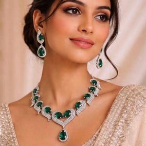 May include: A jewelry set consisting of a necklace and matching drop earrings. The necklace features a central heart-shaped motif with a dangling pear-cut green stone, surrounded by smaller green stones and clear crystals set in silver-toned metal.