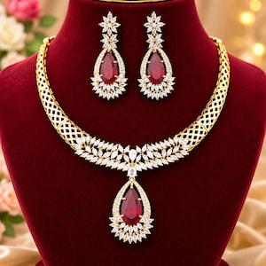 Könnte beinhalten: Ein goldfarbenes Halsketten- und Ohrring-Set mit einem gitterartigen Collier, verziert mit klaren Steinen im Marquise-Schliff und einem zentralen, tropfenförmigen tiefroten Stein. Die passenden Ohrhänger spiegeln das Tropfendesign wider.