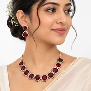 May include: A jewellery set consisting of a necklace and matching earrings featuring deep red, faceted, rounded stones. Each stone is framed by a thin border of small white beads and set in a metallic base, creating a cohesive and ornate appearance.