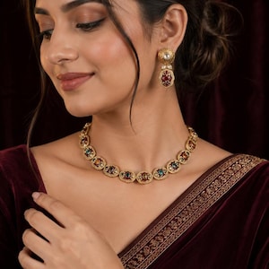 May include: A gold-toned necklace and matching drop earrings set featuring oval-shaped links with intricate metalwork. Each link contains a central gemstone in shades of deep red, emerald green, and amber, accented by small white pearls.
