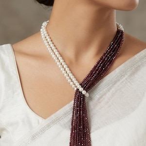 May include: A multi-strand necklace featuring a combination of white pearl beads and deep burgundy faceted crystal beads, arranged in a layered lariat style. The set includes a matching single white pearl drop earring with a silver-tone hook.