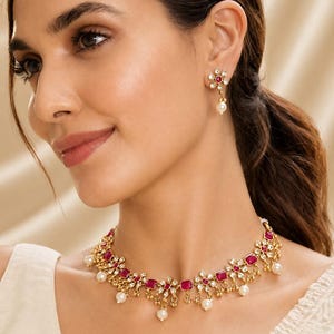 May include: A gold-toned necklace and matching stud earrings set featuring square-cut magenta stones, clear crystal accents arranged in a floral pattern, and small dangling white pearls along the lower edge of the necklace and earring drops.