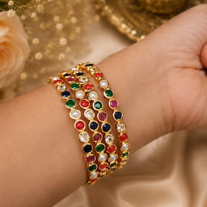 May include: Gold bangles adorned with colourful gemstones and pearls. The bracelets feature red, green, blue, and white stones, creating a vibrant and elegant look. The bangles are stacked on a wrist, showcasing the jewellery's design.