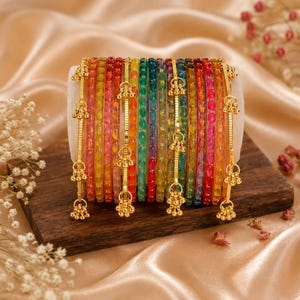 Peut inclure: Une collection de bracelets colorés dans diverses teintes, dont le rouge, le jaune, le vert et le bleu. Chaque bracelet est orné d'éléments décoratifs dorés. Les bracelets sont disposés sur une surface en bois.