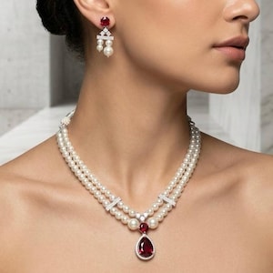 Pode incluir: Um colar de pérolas de duas voltas com um pingente central de pedra vermelha em forma de gota e brincos pendentes a combinar. Cada peça é feita de metal prateado, pequenas pedras claras e pérolas brancas, formando um conjunto coordenado.